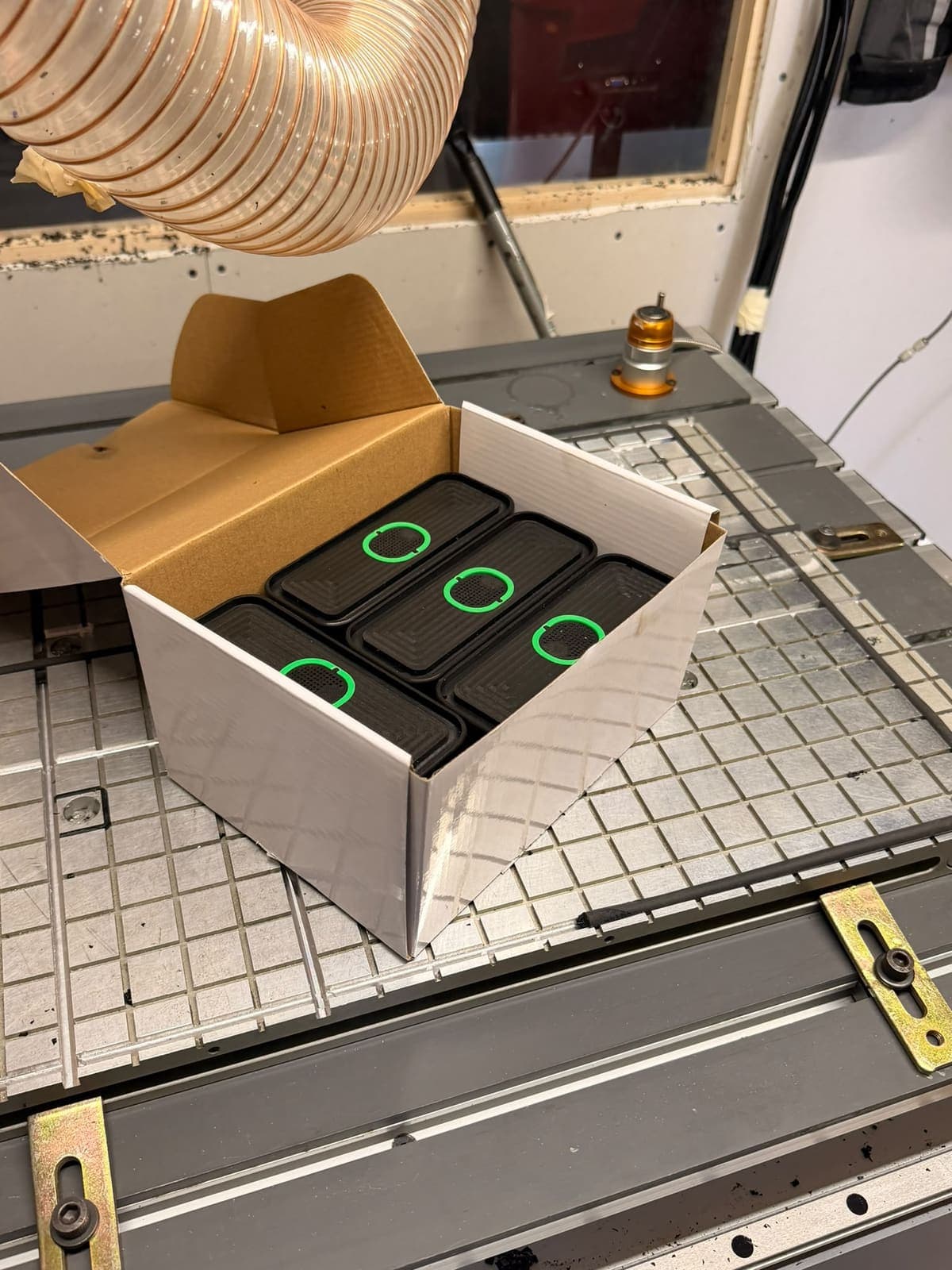 A box of six black vacuum pods with green rings sitting on the grid bed of a CNC machine